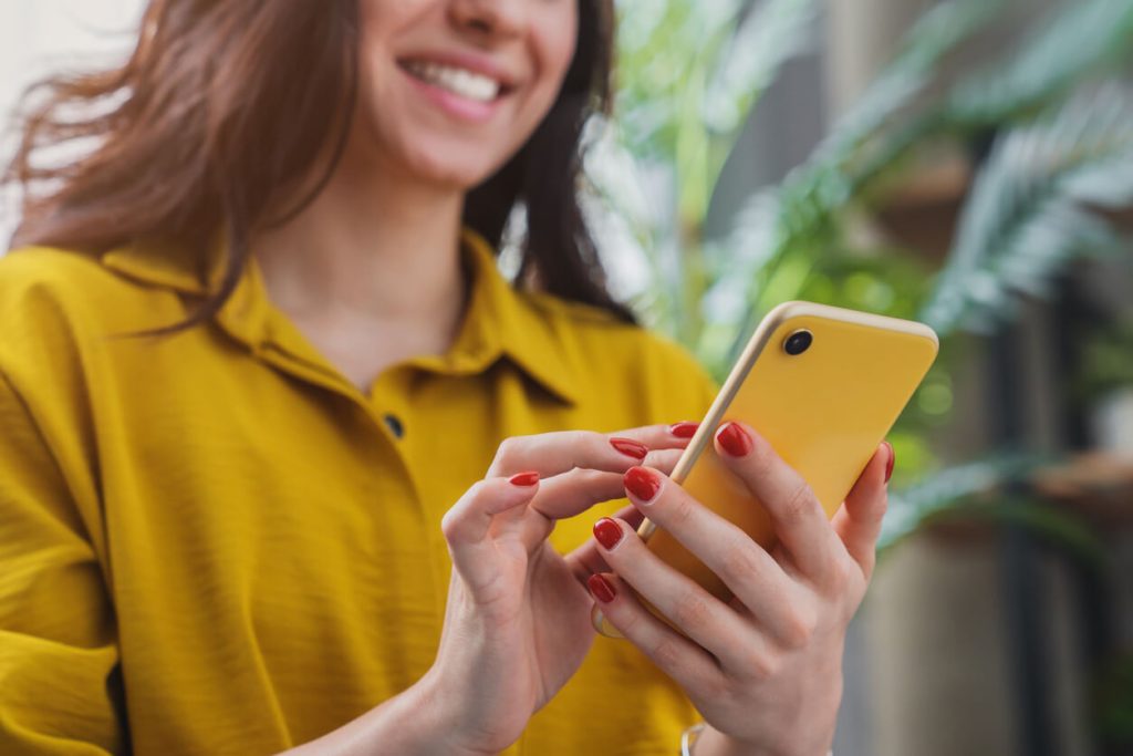 A cheerful young woman using her phone as she walks. She is wearing a yellow shirt which matches her yellow phone case