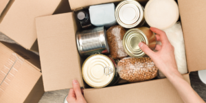 A view of a box filled with canned and nonperishable goods from above. You can see a person's hands adding a can into the box.